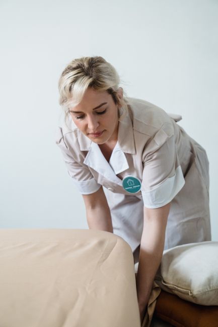 A female cleaner with blonde hair tied back, dressed in a white uniform, is performing surface cleaning on a beige upholstered sofa in a well-lit room with plain white walls. She appears to be focusin